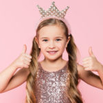 cheerful little girl in dress and crown showing thumbs up e1618726054415