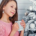 girl child near a christmas tree with a cup in hand e1619076183489